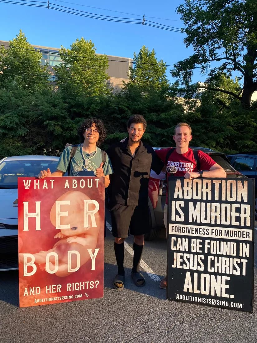 Three abolitionists holding signs reading "What about her body and her rights?" and "Abortion is murder — forgiveness for murder can be found in Jesus Christ alone."