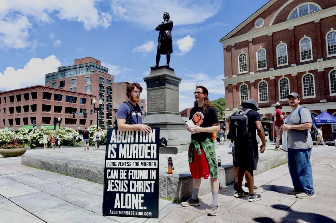 An abolitionist holding an "Abortion is murder" sign beside a statue of Samuel Adams while others speak with passers-by.