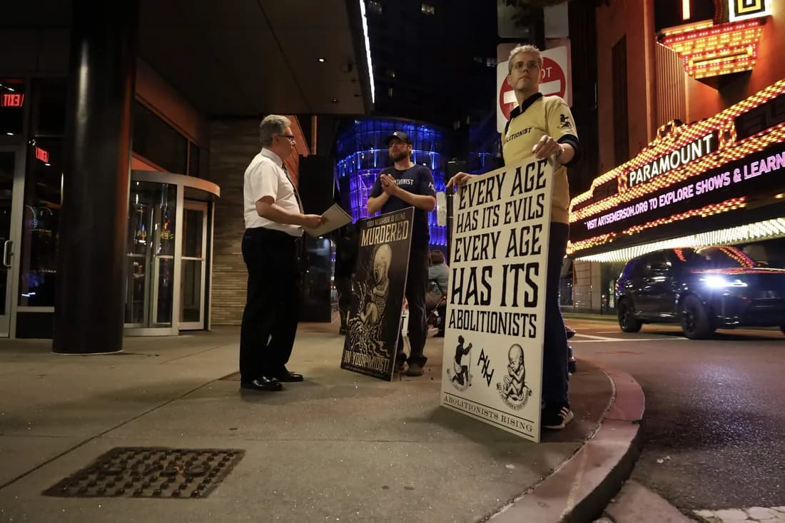 Two abolitionists talking with a man on a city street at night, holding signs reading "Your neighbour is being murdered in your midst" and "Every age has its evils, every age has its abolitionists."