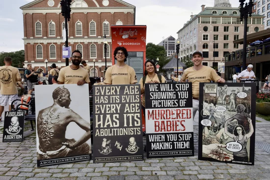 Four abolitionists in ABOLITIONIST shirts standing behind a row of signs comparing abortion to historic chattel slavery.